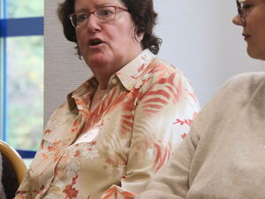 Frau mit braunen lockigen Haaren, rötlicher Brille und musterten hellen Bluse sitzt auf Stuhl. Hände im Schoß gefaltet. Sie spricht.