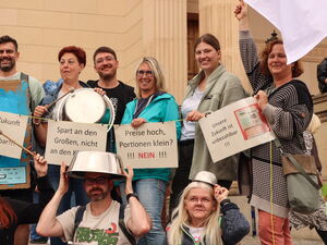 15 Menschen. 6 Sitzen und Knien vor den stehenden Personen. Die erste Reihe hat große Kochtopfe auf dem Kopf. Die hintere Reihe hält Protest-Schilder in den Händen.
