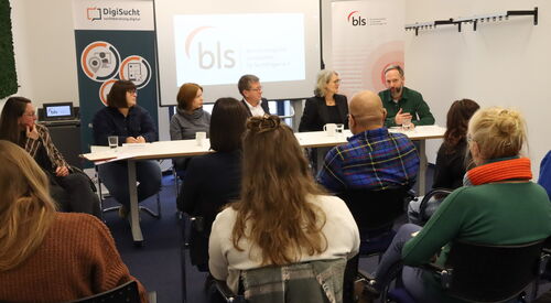 Vier Personen an einem langen Tisch. Drei Frauen und Zwei Männer nehmen an der Diskussion teil.. Zuschauende / Zuhörende sitzen davor.