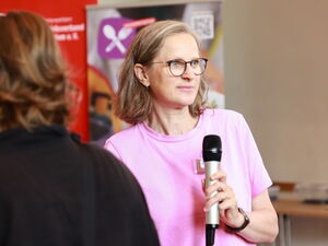Frau hält Mikrofon in der Hand und steht im Raum. Frau trägt Brille und ein rosa T-Shirt, sie hält ein Mikrofon in der Hand.