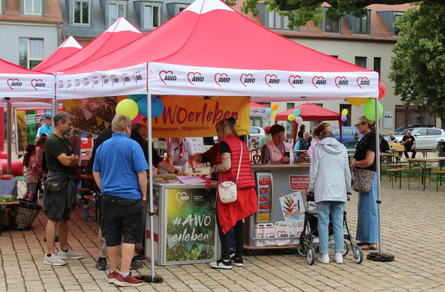 Rote AWO Pavillons: gelbes Banner: "AWO erleben"   Menschen am Stand  die sich informieren.