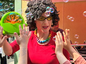 Frau mit schwarzer lockiger Perücke, Sonnenbrille, bunter Perlenkenne und rotem Oberteil hält Seifenblasenmaschine in der Hand. Kinderhände greifen nach den Seifenblasen.
