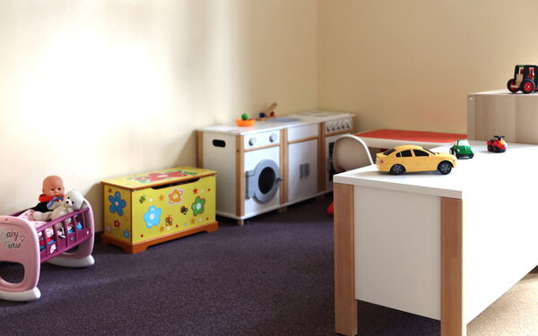 In der Zimmerecke ein Kinderherd, Spüle und Waschmaschine. Daneben eine gelbe Truhe mit bunten Blumen drauf.