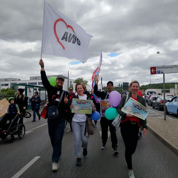 Vier Menschen die auf die Kamera zugehen. Eine AWO Fahne wird erhoben. Zwei Frauen halten kleine Plakate vor sich.
