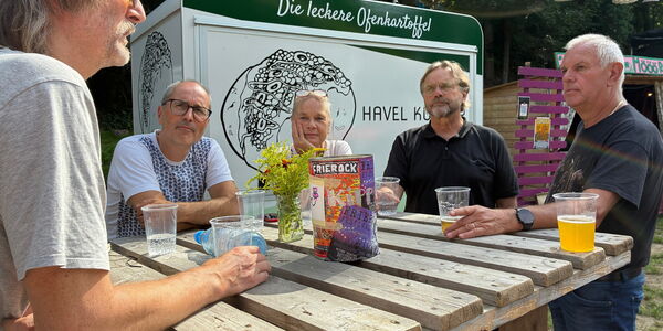 5 Menschen an einem großen Stehtisch aus Holz mit Getränken in lockerer Runde.