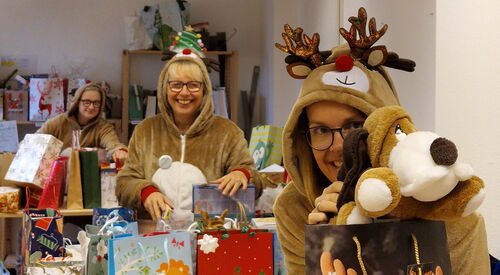 Gefüllte Geschenktüten. Ganz votn mit einem Weihnachtshund drauf. Frau im Rentierkostüm hält einen Plüschhund darüber und lächelt. Dahinter noch eine Frau im Rentierkostüm mit mehreren Geschenktüten. Alle sind sehr fröhlich.
