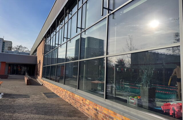 Schwimmhalle mit großem Fenster, ein großes Becken im inneren. Die Sonne spiegelt sich im Fenster.