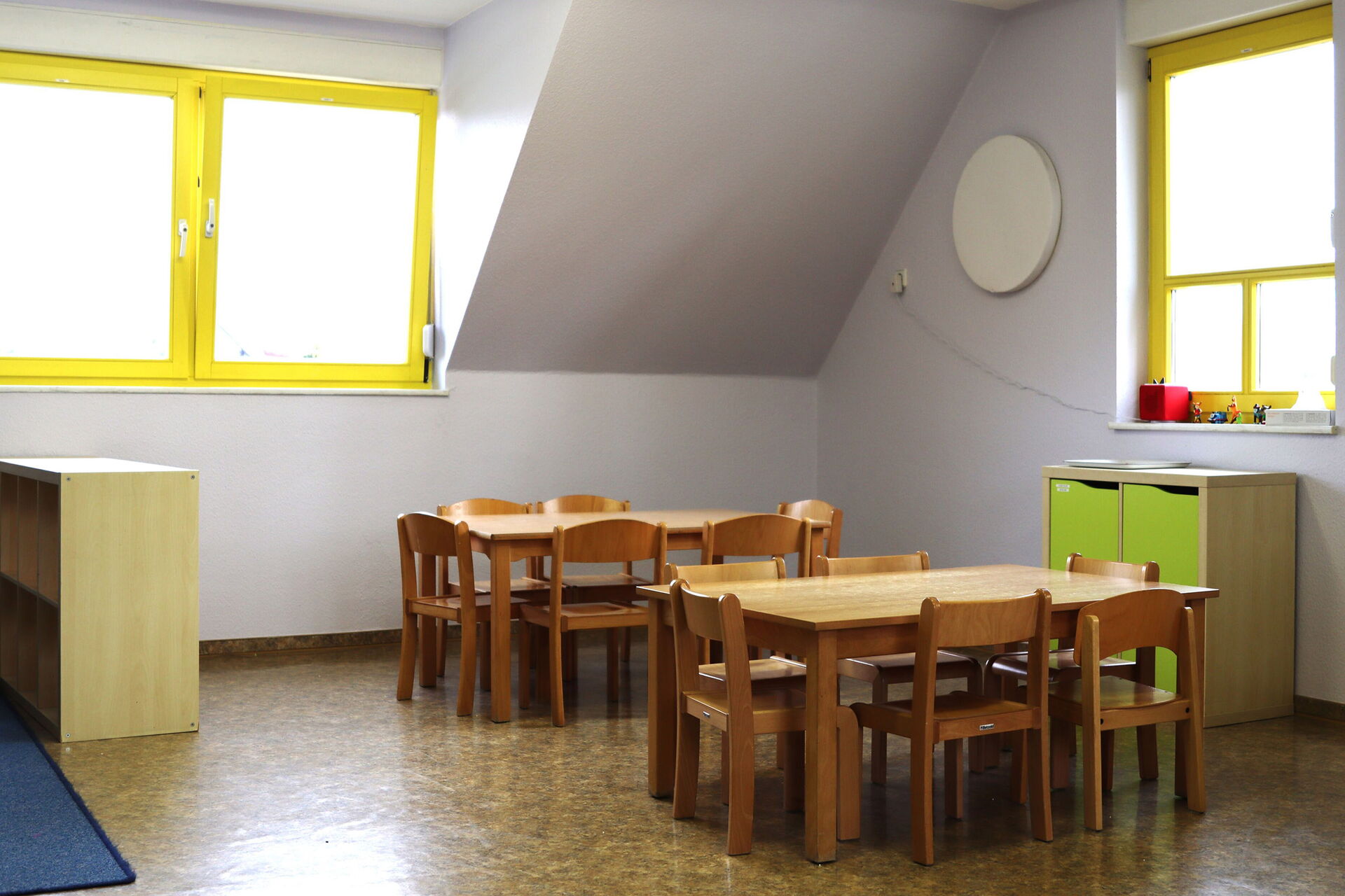 Heller Raum mit zwei kleinen Tischen mit jeweils 6 kleinen Stühlen. Ein kleines Schränkchen steht an der Wand unter einem der Fenster mit gelben Fensterrahme.