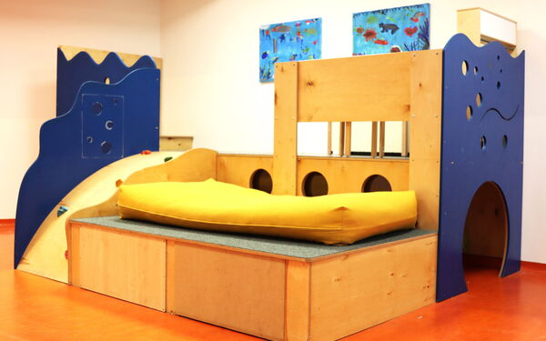 Klettergerüst mit Höhle, Liegefläche mit gelber Matratze, kleiner Rutsche. Es ist aus Holz, einige Elemenze sind blau und Wellenförmig wie das Meer.