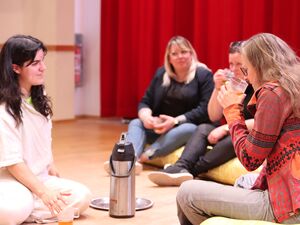 Beim Teetrinken. Personen sitzen auf dem Boden und auf gelben Matratzen und trinken Tee. Eine Thermos Drückkanne steht in der Mitte auf dem Fußboden.