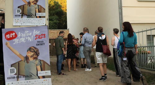 Draußen. Häuser. Große Plakate von Kultur für Jeden an Hauswand. Eine kleine Gruppe Mensch in sommerlicher Kleidung.