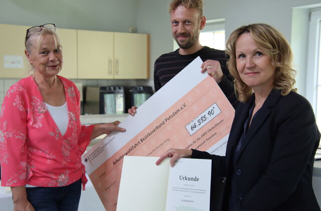 Drei Personen in einem Raum. Blonde Frau und Mann mit Vollbart halten einen großen Scheck über 64.385,90 Euro in den Händen. Frau mit braunen gewellten schulterlangen Haaren und dunklem Blazer hält eine Urkunde in die Kamera.
