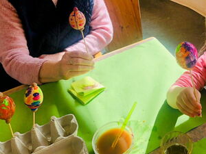 Beim Eier bemalen. Bunte Eier sind zum Trocknen auf Spieße gespießt. Eine Frau und ein Kind halten ihre Eier auf dem Spieß.