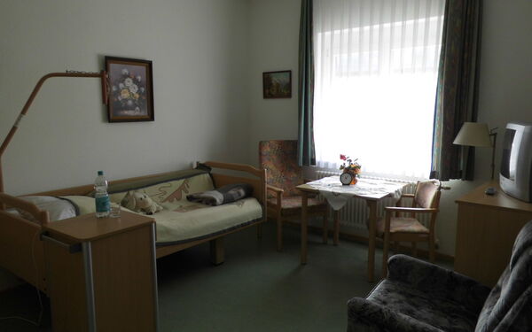 Bewohnerzommer, gemütlich eingerichtet mit Bett, Sessel, Tisch und 2 Stühlen am Fenster.