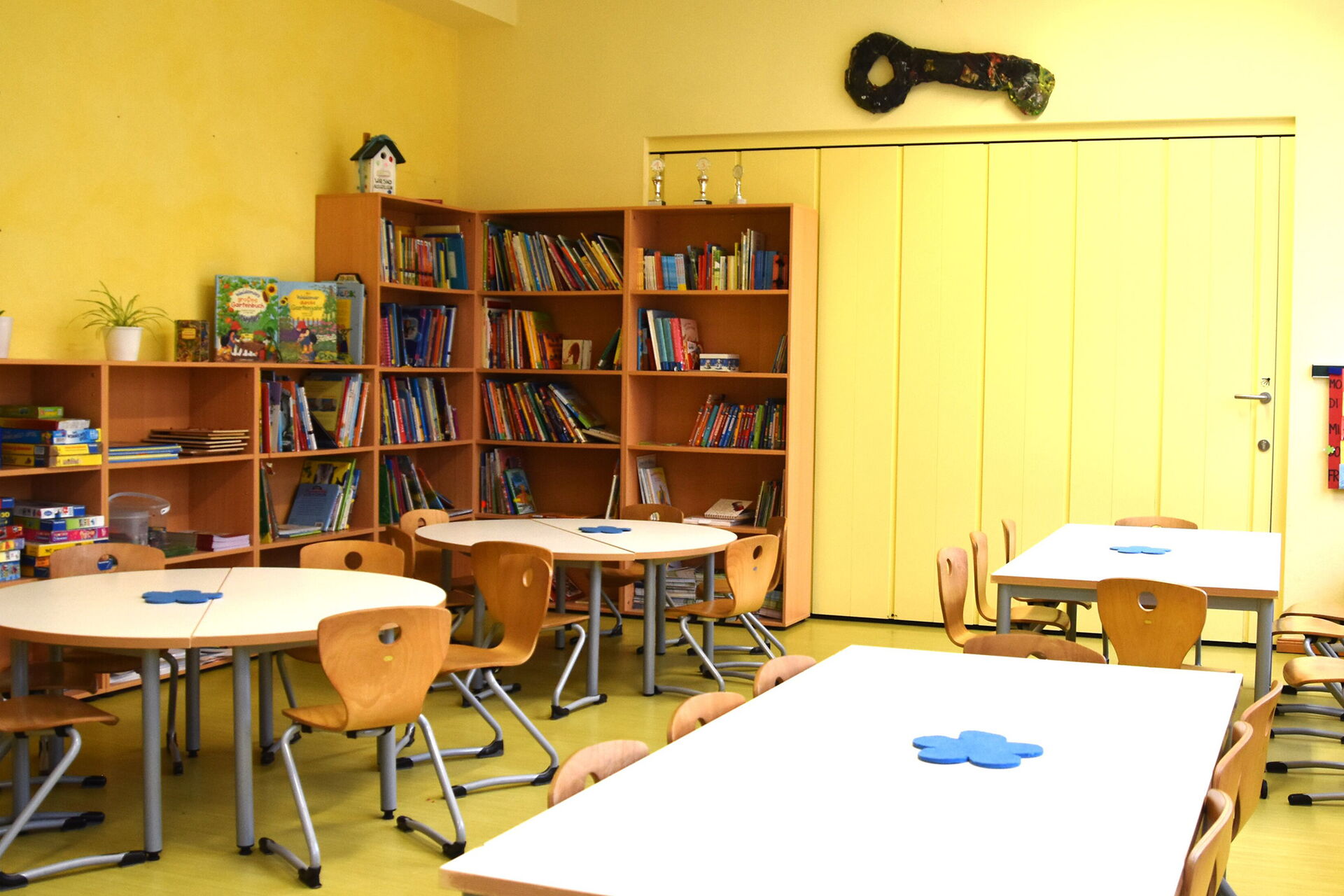 Runde und rechteckige Tische mit Stühlen und große Regale in der Ecke und an der linken Wand. In den Regalen Brettspiele und Bücher.