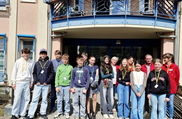 Gruppenbild vor dem Eingang eines Gebäudes. 15 Personen. 4 Erwachsene in roten AWO Jacken und Schüler.