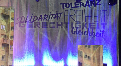 Bühnenvorhang mit Text: Toleranz, Solidarität, Freiheit, Gerechtigkeit, Gleichheit. Dahinter wird die Kulisse des Theaterstücks sichtbar. Ein Häuserblock.