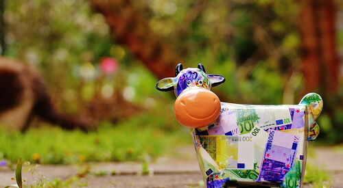 Bunte Kuh-Spardose mit aufgedruckten Euro-Geldscheinen in verschiedenen Werten steht auf einem Gartenweg vor unscharfem Naturhintergrund.