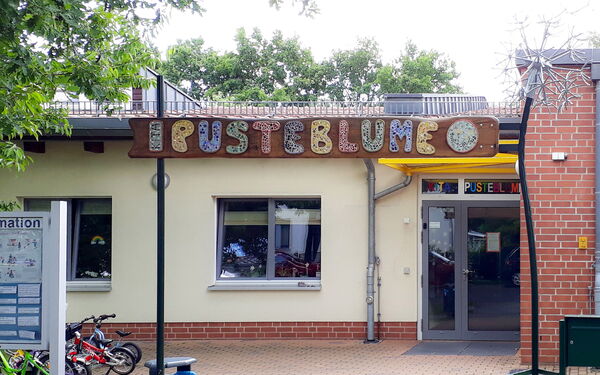 Eingang einer Kita. In großen farbigen Buchstaben über dem Eingang steht das Wort: PUSTEBLUME