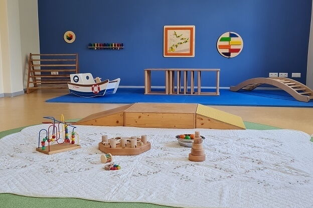 Im Vordergrund: Decke mit Spielzeug für Babys. Im Hintergrund Kletterelemente und eine blaue Wand mit Bild.