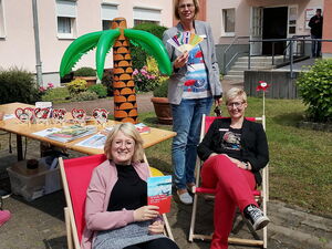 Ein Haus, im Vordergrund zwei Frauen sitzen in roten Sonnenstühlen unter einer aufblasbaren Gummipalme. Eine Frau steht hinter ihnen mit einem Fächer in der Hand.