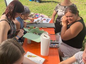 Am einem Holztisch sitzen 6 Menschen. Ein Junge im T-Shirt bekommt ein Henna Tattoo auf die Hand gemalt.