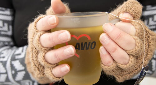 Teetasse mit AWO-Logo. Tasse wird mit zwei Händen in fingerlosen braunen Handschuhen gehalten.