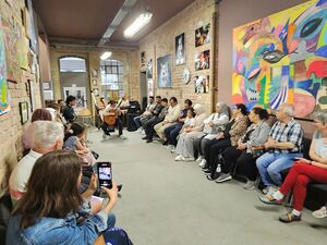 Langgezogener Raum, Gäste sitzen zu beiden Seiten auf Stühlen. An den Wänden sind große Bilder zum Teil Abstrakt. Ganz am Ende des Raums sitzt eine Frau und spielt Gitarre.