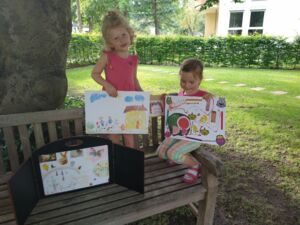 Zwei Mädchen in rosa Sommerkleidung. Sie befinden sich auf einer Bank unter einem Baum. Sie halten Bilder von Kindern in die Kamera. Eines zeigt ein gelbes Haus und eine kleinee Familie. Auf dem anderen Bild ist eine Melone.