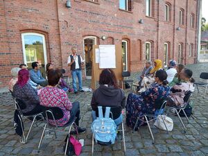 Draußen, Kopfsteinpflaster, Gebäude. In einen Kreis sitzen 13 Menschen auf Stühlen. Ein Mann steht neben einem Flipchart und erklärt etwas.