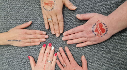 5 Hände von verschiedenen Menschen im Kreis Fin, Auf den Händen sind Stempel: AWO GEGEN RASSISMUS Herz mit Schärpe | AWO GEGEN RASSISMUS MUTIG in der Mitte.