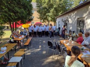 Draußen. Tische mit Kaffeetassen an denen weinige Menschen sitzen. Weiter hinten stehen viele Menschen in weißen T-Shirts.