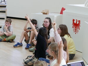 Kinder sitzen auf dem Fußboden im Plenarsaal. Einige melden sich.