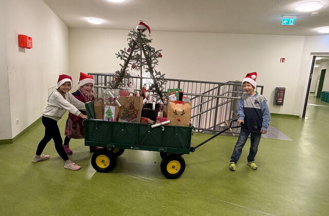 Ein Handwagen beladen mit Geschenken wir von zwei Kindern geschoben ein Junge zieht vorne am Griff. Sie sind in einem Gebäude auf einem Flur und tragen rote Weihnachtsmützen.