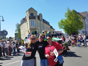 Festumzug. 3 Frauen mit AWO T-Shirt stehen im Vordergrund und lächeln in der Kamera. Dahinter ein geschückter Traktor. Die Straßen gesäumt von Menschen.