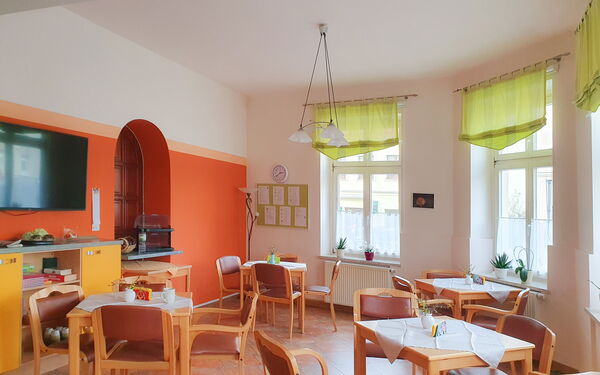 Speisesaal. Halbrunder Raum mit vier Tischen mit jeweils vier Stühlen. Heller freundlicher Raum, links: ein kleines Regal darüber ein Fernseher an einer Wand die orange und oben weiß ist.