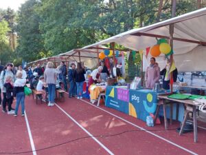 Bunte Marktstände, mit Luftballons und Wimpeln dekoriert. Viele Menschen von jung bis alt sehen sich die Stände an oder stehen beieinander.