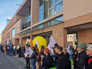 Viele Menschen mit Schildern und auch AWO Fahnen die hochgehalten werden. Auf einem Gehweg vor einem langen großen Gebäude mit vielen Fenstern.