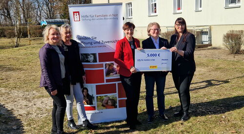 Draußen, Sonne scheint. Mittig ein Roll-Up von „Hilfe für Familien in Not“. Links davon zwei lächelnde Frauen. Rechts davon drei Frauen halten Scheck in die Kamera.