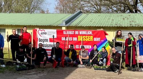 15 Menschen unterschiedler Nationen mit Pflanzen und Gartengeräten, Besen, Harke und Schaufeln. Die Menschen stehen und hocken vor dem AWO Banner