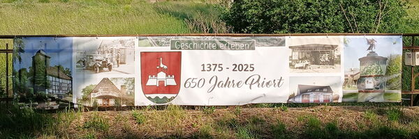 Ein Banner befestigt an einem Zaun mit historischen Fotos und Text: Geschichte erleben       1375 - 2025    650 Jahre Priort.