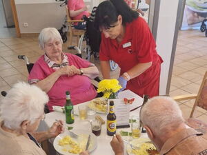 Mittag. Tisch mit drei älteren Personen, die Mittag essen. Eine Betreuerinn in roter Kleidung schneidet einer Seniorin das essen.