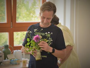 Mann mit schwrzen T-Shirt mit Aufdruck hält ein Glas mit Wasser in das er einen kleinen Blumenstrauß steckt. Hinter ihm ein Tisch mit Kannen, Tee und Zuckerdose.