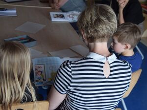 Eine Frau, ein Mädchen und ein Junge sehen sich ein Kinderbuch an. Sie sind von hinten fotografiert. Sie sitzen an einem Tisch. Mädchen mit langen blonden Haaren, Frau mit Kurzhaarfrisur, Nacken frei und trägt ein gestreiftes T-Shirt.