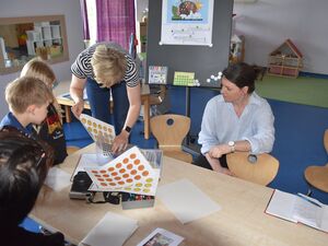 Zwei Kinder und drei Frauen am Tisch. Aufkleber von gelben und orangenen Smileys liegen auf dem Tisch. Frau mit kurzen blonden Haaren kramt noch mehr Smileys hervor. Frau mit schwarzen Haaren hockt am Tisch und stützt sich mit einem Arm auf Kinderstuhl.