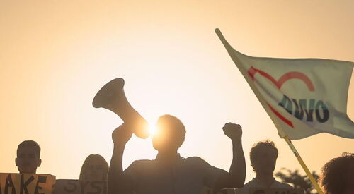 eine Gruppe Menschen demonstriert, eine AWO-Flagge schwingt im Wind. Im Hintergrund geht die Sonne unter.