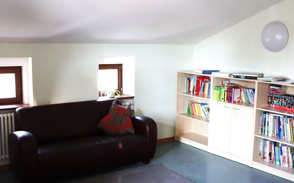 Aufenthaltsraum. Dachgeschoss mit Dachschräge, kleine Fenster. Eine schwarze Couch vor den Fenstern. Regal an der Wand stehend mit Büchern.