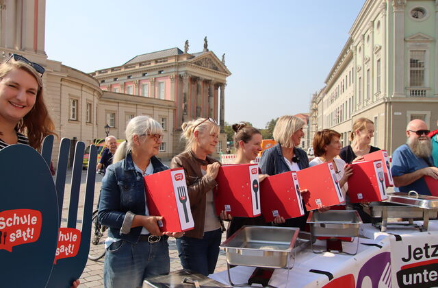 Auf dem Alten Markt in Potsdan. Die Sonne scheint. 6 Frauen und ein Mann stehen in einer Reihe und halten rote volle Aktenordner vor sich die das Logo: "Schule SATT" tragen.