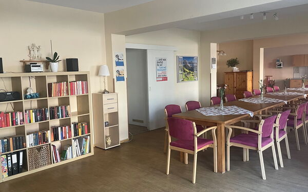 am linken Bildrand steht ein ausreichend gefülltes Bücherregal, darauf steht eine Musikanlage. rechts davon steht in der Mitte des großen Raumes eine lange Tischtafel mit Stühlen.