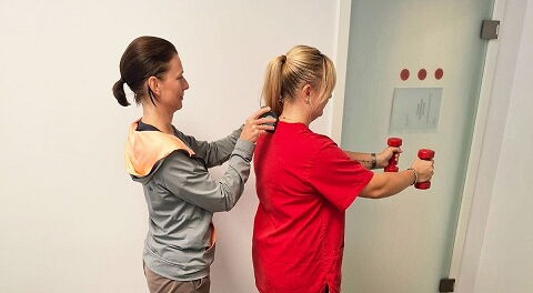 2 Frauen stehen hintereinander, die vordere Person trägt ein rotes T-Shirt und hält zwei rote Hanteln mit ausgestreckten Armen, hinter ihr steht die Therapeutin, sie trägt eine graue Jacke und hat braune Haare, sie korrigiert die Haltung am Rücken.